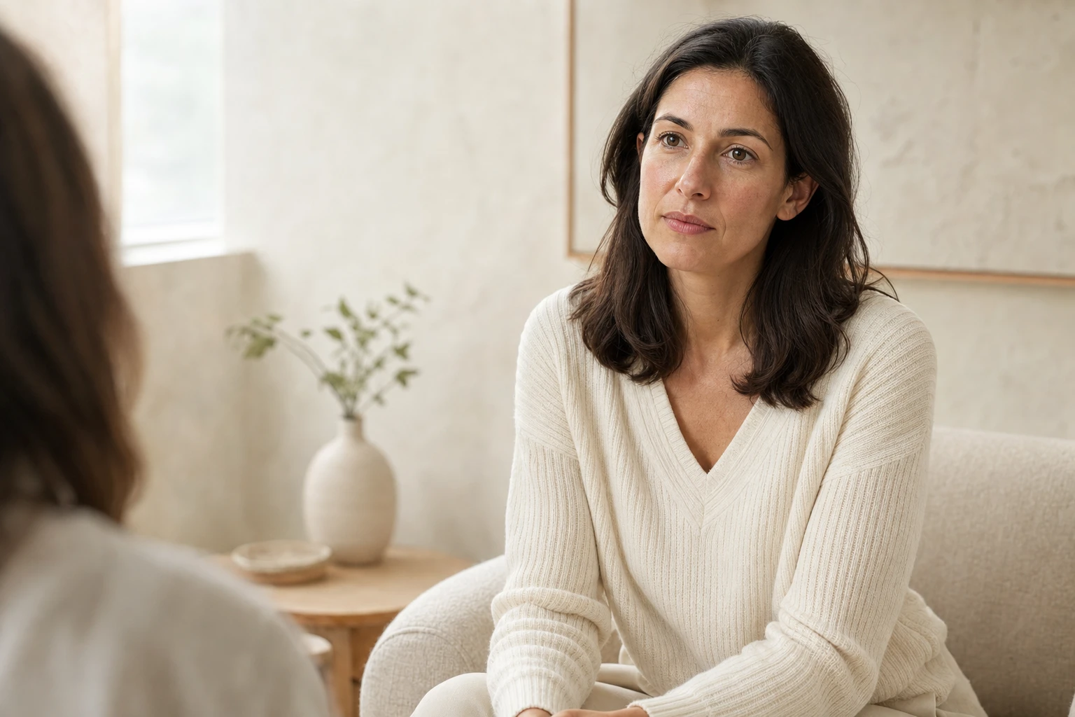 An adult woman with a calm expression listens to the person she is talking to during a discussion, in a bright room with ivory tones.