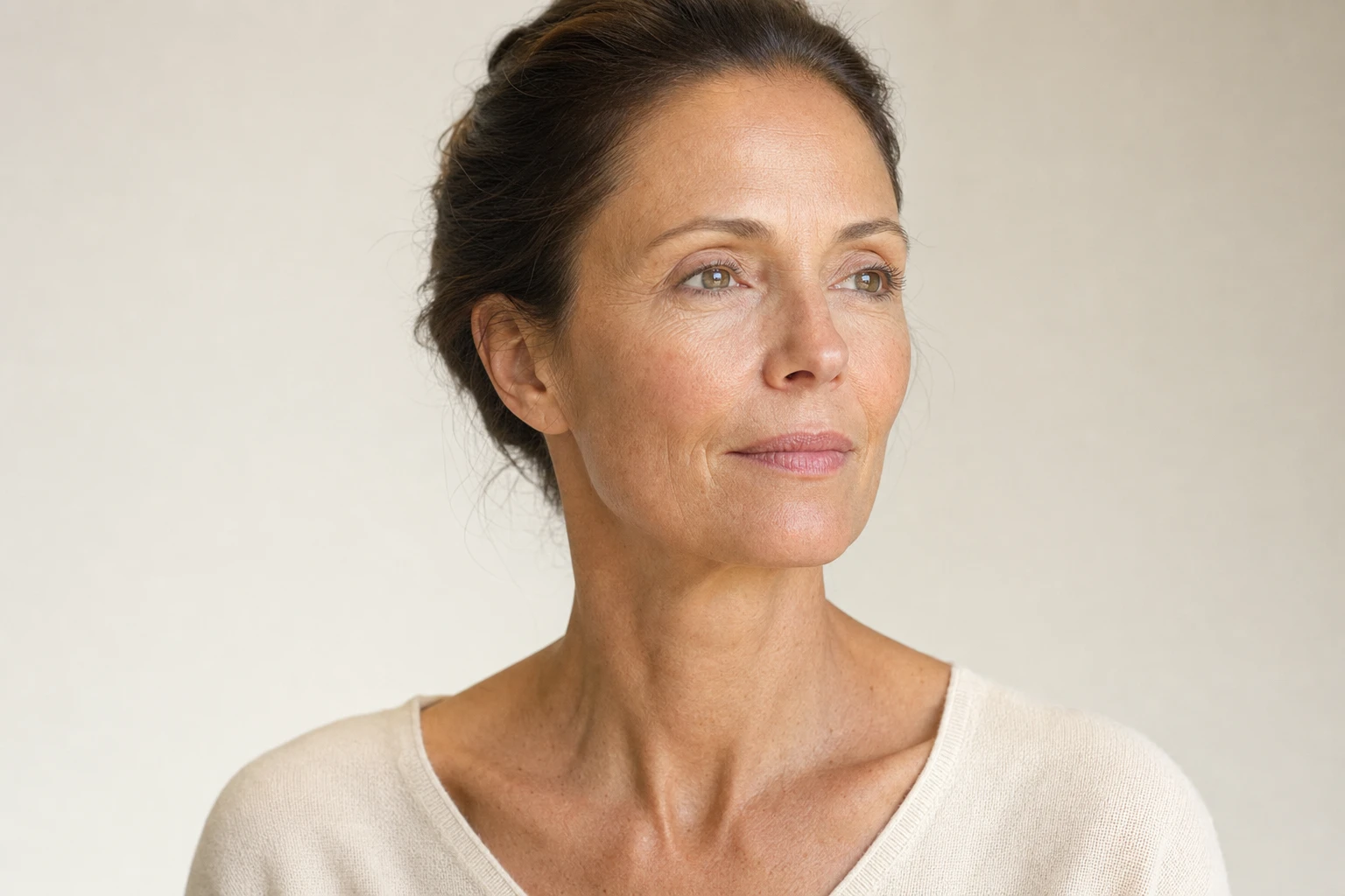 Milieu du visage d'une femme fictive vu de trois quarts, pommettes et joues éclairées par une lumière douce latérale, peau au naturel sans retouche, sur fond clair et sobre.