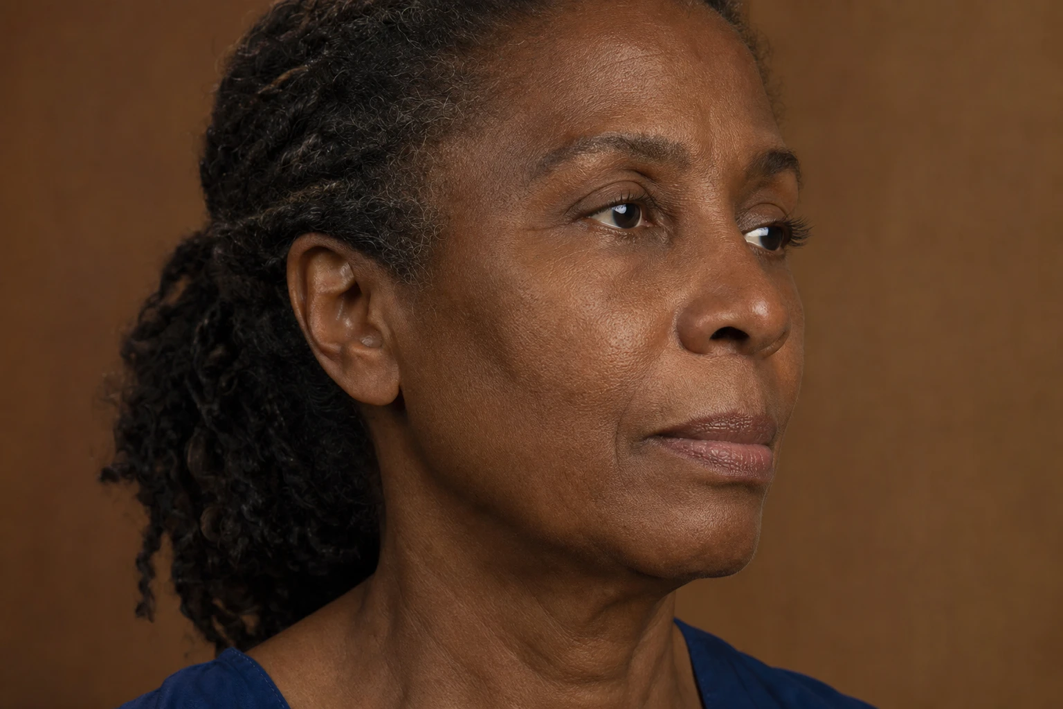 Three-quarter view of the face of a fictional woman of about sixty with dark skin: temple, cheekbone and facial contour visible, with natural texture and folds, against a warm brown background.
