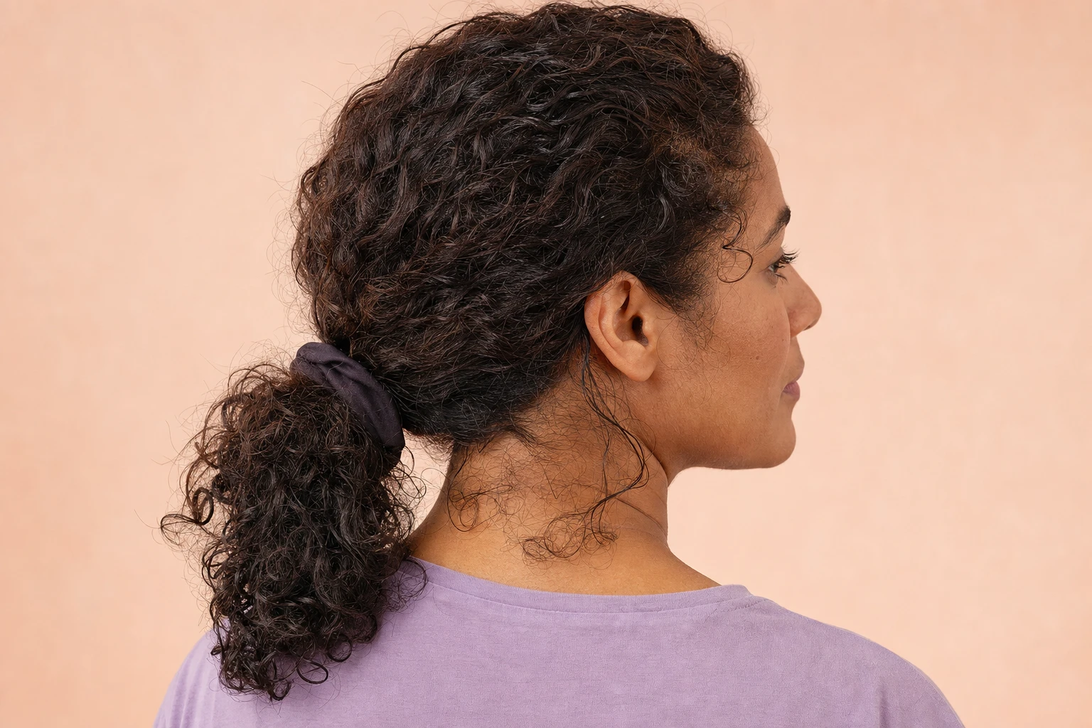 Three-quarter rear view of a woman with curly hair tied low, with loose strands at the nape of her neck.