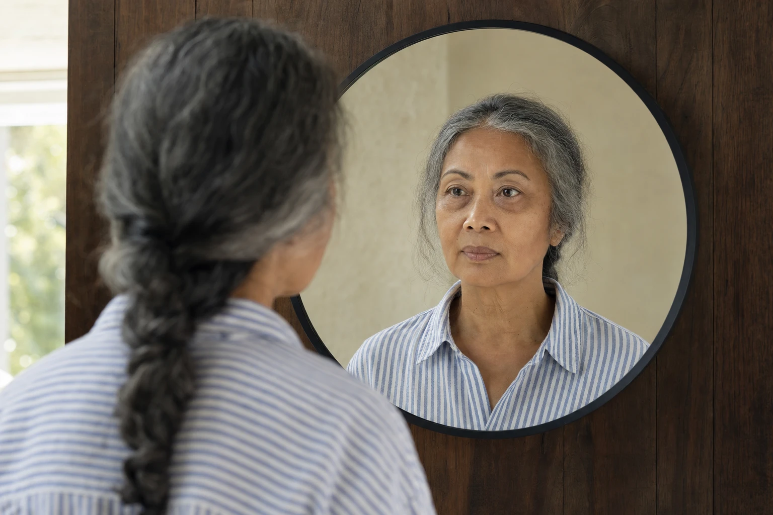 Woman with long braided grey hair looking in a round mirror at the area under her eyes, which is slightly dark and a little puffy.