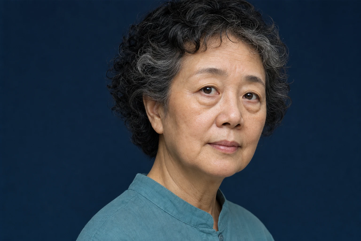 Older woman with curly grey and black hair, in three-quarter view, with a calm expression and slight bags under both eyes.