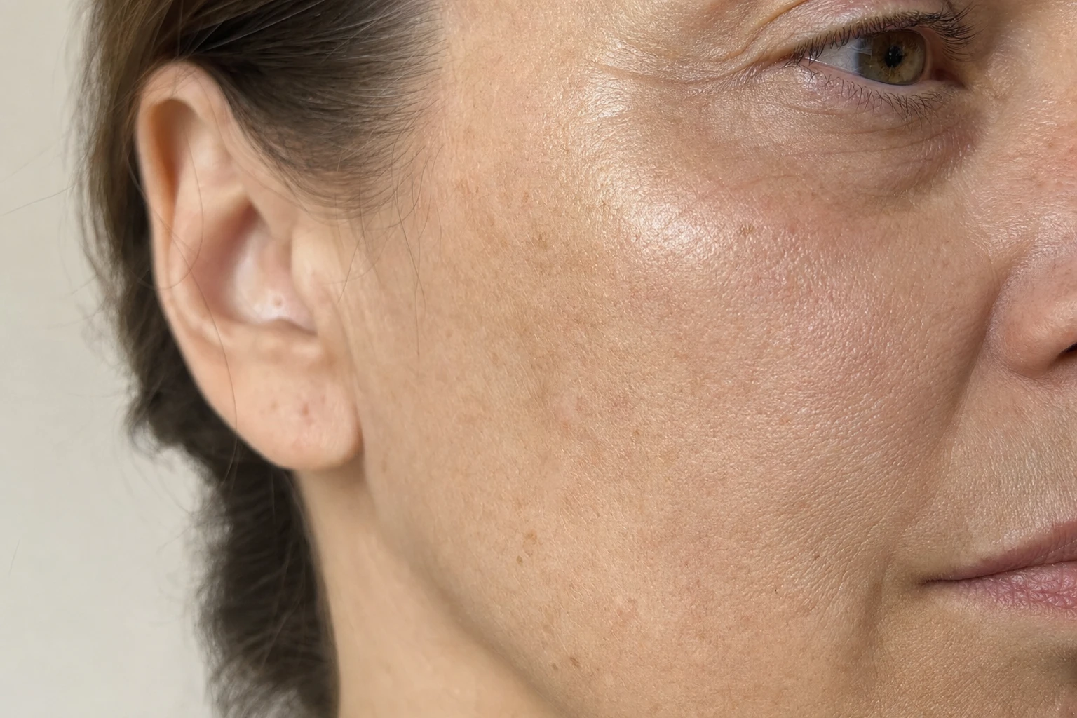 Gros plan sur la joue et la pommette d'une femme fictive, peau au naturel avec pores et ridules visibles, éclairée d'une lumière douce.