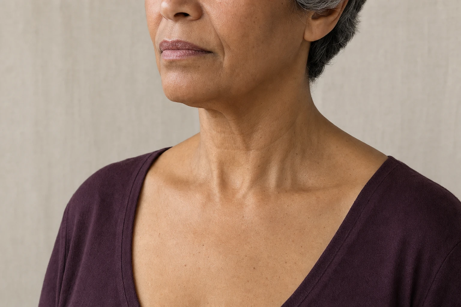 Bas du visage, cou et haut du décolleté d’une femme à la peau mate, de trois quarts, portant un haut prune sur fond lin.
